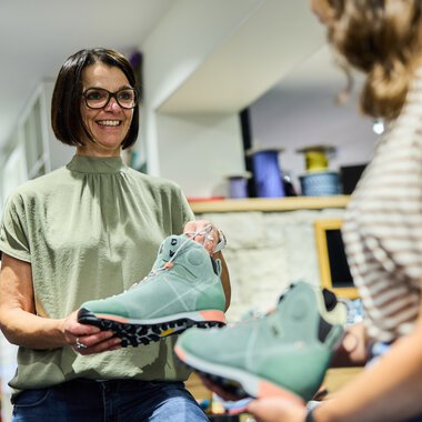 Woman with glasses and short hair holding a pair of blue hiking shoes and talking to a customer in the store. | © Kleinwalsertal Tourismus eGen • Photographer: Oliver Farys