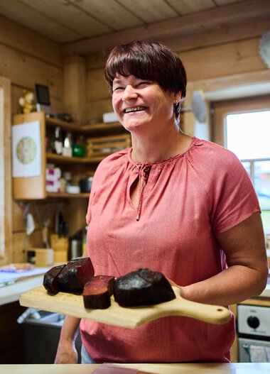 Woman in a wooden cabin kitchen, smiling and holding a cheese slice on a wooden board. | © Kleinwalsertal Tourismus eGen • Photographer: Oliver Farys