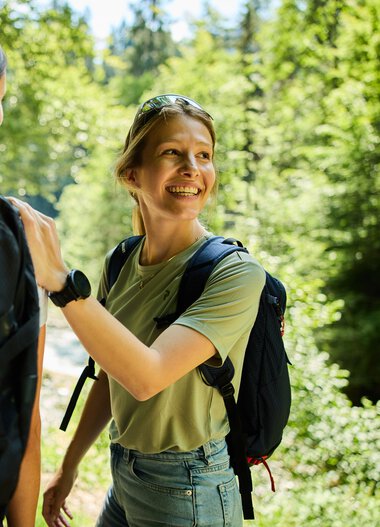 Zwei Frauen mit Rucksack wandern in einem grünen Wald und unterhalten sich fröhlich. | © Kleinwalsertal Tourismus eGen • Fotograf: Oliver Farys