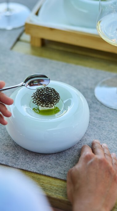 Ein gedeckter Tisch mit einer modernen weißen Platte, auf der ein Teller mit einem kleinen Gericht und Stäbchen liegt. Es gibt auch ein Glas Weißwein, ein Wasserglas mit Minzblättern und eine silberne Gabel. Der Tisch hat eine Holzoptik und eine braune Decke mit Jagdmotiv. | © Kleinwalsertal Tourismus eGen • Fotograf: Oliver Farys