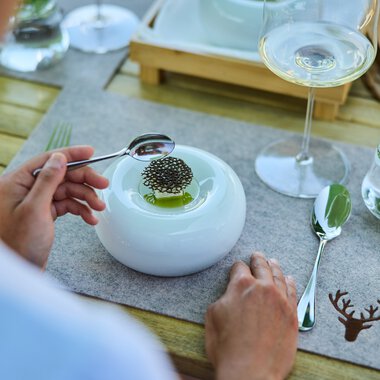 Ein gedeckter Tisch mit einer modernen weißen Platte, auf der ein Teller mit einem kleinen Gericht und Stäbchen liegt. Es gibt auch ein Glas Weißwein, ein Wasserglas mit Minzblättern und eine silberne Gabel. Der Tisch hat eine Holzoptik und eine braune Decke mit Jagdmotiv. | © Kleinwalsertal Tourismus eGen • Fotograf: Oliver Farys