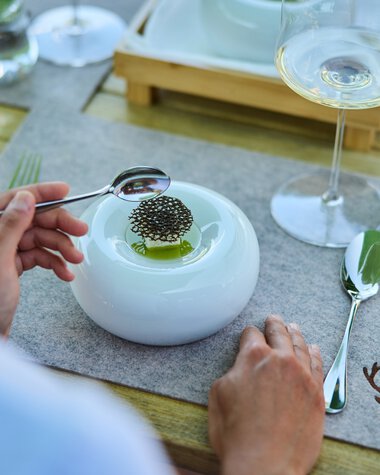 A table setting featuring a modern white plate with a small dish and chopsticks, a glass of white wine, a water glass with mint leaves, and utensils on a wooden table with a nature-themed tablecloth. | © Kleinwalsertal Tourismus eGen • Photographer: Oliver Farys