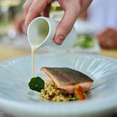 Ein Koch gießt cremige Sauce über ein Gericht mit Fisch, serviert auf einem weißen Teller bei einem kulinarischen Event. | © Kleinwalsertal Tourismus eGen • Fotograf: Oliver Farys