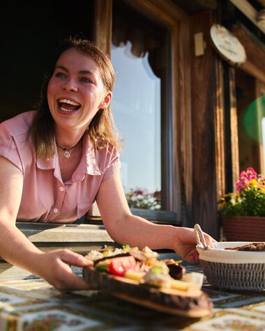 A smiling young woman stands at a window of a rustic mountain hut, holding a served meal and enjoying the summer atmosphere in Kleinwalsertal. | © Kleinwalsertal Tourismus eGen • Photographer: Oliver Farys