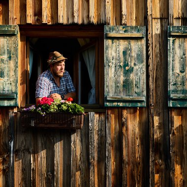 Mann schaut aus einem Fenster einer rustikalen Holzfassade mit grünen Schiebefenstern, umgeben von Blumen. | © Kleinwalsertal Tourismus eGen • Fotograf: Oliver Farys