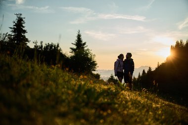 Zwei Menschen stehen beim Sonnenaufgang in einer Landschaft mit Bäumen und Hügeln und genießen die Natur im Kleinwalsertal. | © Kleinwalsertal Tourismus eGen • Fotograf: Oliver Farys