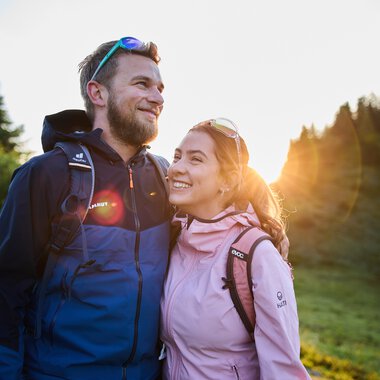 A smiling couple hiking during summer sunrise in a forested area. | © Kleinwalsertal Tourismus eGen • Photographer: Oliver Farys
