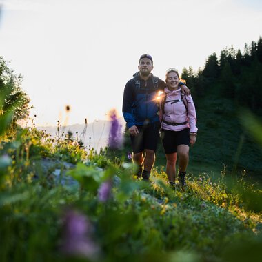 Ein Paar wandert im Frühling im Kleinwalsertal bei Sonnenaufgang, umgeben von Blumen und grünen Bergen. | © Kleinwalsertal Tourismus eGen • Fotograf: Oliver Farys