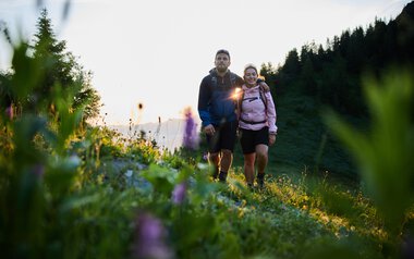 Ein Paar wandert im Frühling im Kleinwalsertal bei Sonnenaufgang, umgeben von Blumen und grünen Bergen. | © Kleinwalsertal Tourismus eGen • Fotograf: Oliver Farys