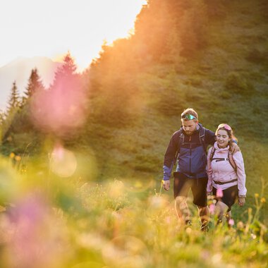 Paar wandert in den frühen Morgenstunden durch eine Berglandschaft im Kleinwalsertal bei Sonnenaufgang, umgeben von grünen Wiesen und Bäumen. | © Kleinwalsertal Tourismus eGen • Fotograf: Oliver Farys