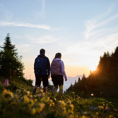 A couple hiking during sunrise in the Kleinwalsertal, surrounded by trees and flowers, with the sky and rising sun in the background. | © Kleinwalsertal Tourismus eGen • Photographer: Oliver Farys
