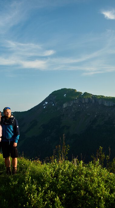 Ein Paar wandert bei Sonnenaufgang in den Bergen des Kleinwalsertals, umgeben von grüner Landschaft. | © Kleinwalsertal Tourismus eGen • Fotograf: Oliver Farys