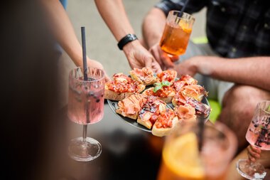 Menschen bei einem Sommer-Event im Kleinwalsertal, die Pizza servieren und Cocktails trinken. | © Kleinwalsertal Tourismus eGen • Fotograf: Oliver Farys