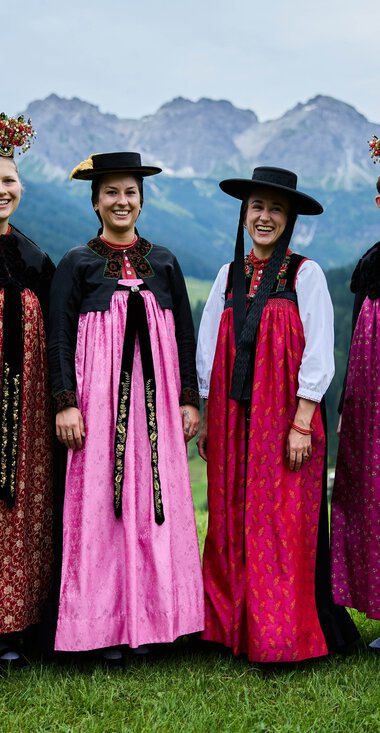 Vier Frauen in traditioneller Tracht stehen auf einer grünen Wiese vor einer bergigen Landschaft mit Wäldern und Bergen im Hintergrund. | © Kleinwalsertal Tourismus eGen • Fotograf: Oliver Farys