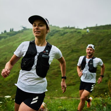 Zwei Trailrunner, eine Frau und ein Mann, laufen auf einem grünen Hügel in Kleinwalsertal bei sommerlichem Wetter. Beide tragen Laufkleidung und Tragegurte. | © Kleinwalsertal Tourismus eGen • Fotograf: Oliver Farys