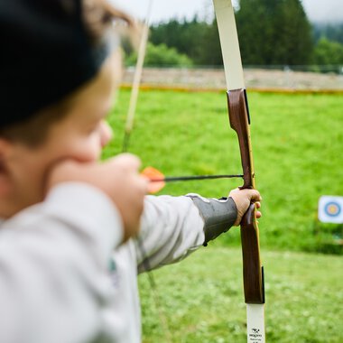A child practicing archery at a wilderness school in Kleinwalsertal, aiming at a target in the background, surrounded by green nature. | © Kleinwalsertal Tourismus eGen • Photographer: Oliver Farys