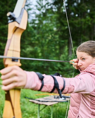 Mädchen beim Bogenschießen in der Wildnisschule im Kleinwalsertal, umgeben von grüner Natur. | © Kleinwalsertal Tourismus eGen • Fotograf: Oliver Farys