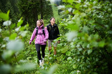 A man and a woman hiking together through a lush green forest in summer, smiling and enjoying nature. | © Kleinwalsertal Tourismus eGen • Photographer: Oliver Farys
