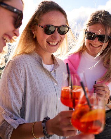 Three women smiling and clinking glasses with Aperol Spritz at a summer lounge in Kleinwalsertal. | © Kleinwalsertal Tourismus eGen • Photographer: Oliver Farys