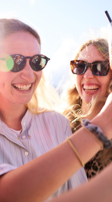 Three women smiling and toasting with drinks outdoors on a sunny day. | © Kleinwalsertal Tourismus eGen • Photographer: Oliver Farys
