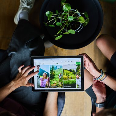 Zwei Personen betrachten Fotos auf einem Tablet, das auf ihren Knien liegt, in einem Büro im Kleinwalsertal. | © Kleinwalsertal Tourismus eGen • Fotograf: Oliver Farys