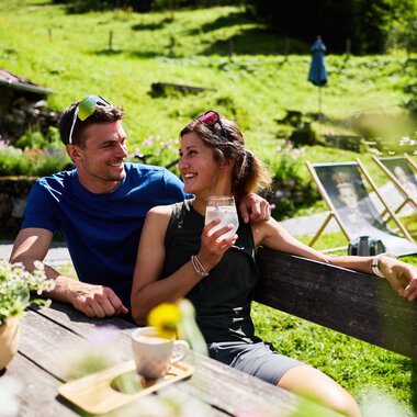 Zwei Freunde sitzen auf einer Bank im Sommer in einem Grünen, lachen und trinken gemeinsam draußen. | © Kleinwalsertal Tourismus eGen • Fotograf: Oliver Farys
