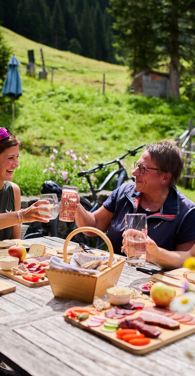 Four friends sitting at a wooden table outdoors in the countryside, enjoying drinks and grilled food on a sunny day in the mountains, with meadows, trees, and a house in the background. | © Kleinwalsertal Tourismus eGen • Photographer: Oliver Farys
