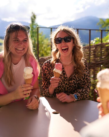 Three women sitting outside at a table enjoying ice cream, laughing and basking in the sun with a mountain landscape in the background. | © Kleinwalsertal Tourismus eGen • Photographer: Oliver Farys