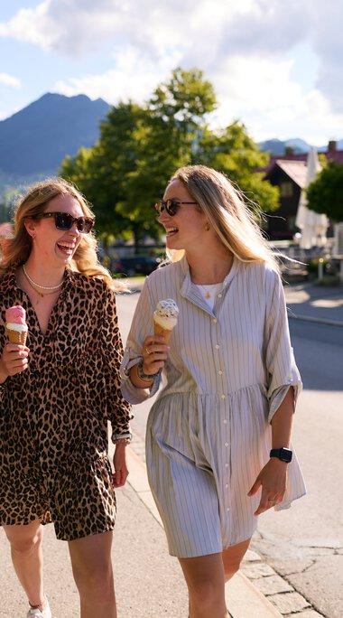 Three young women happily walking in Kleinwalsertal in summer, each holding an ice cream, surrounded by green trees and mountains in the background. | © Kleinwalsertal Tourismus eGen • Photographer: Oliver Farys