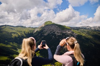 Two women taking photos of a stunning mountain landscape in Kleinwalsertal, surrounded by forests and cloudy sky. | © Kleinwalsertal Tourismus eGen • Photographer: Oliver Farys