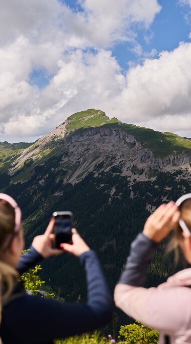 Two women taking photos of a stunning mountain landscape in Kleinwalsertal, surrounded by forests and cloudy sky. | © Kleinwalsertal Tourismus eGen • Photographer: Oliver Farys