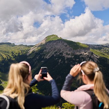 Zwei Frauen fotografieren eine beeindruckende Bergkulisse im Kleinwalsertal, umgeben von Wäldern und bewölktem Himmel. | © Kleinwalsertal Tourismus eGen • Fotograf: Oliver Farys