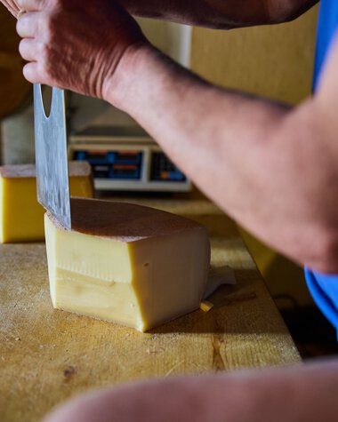 Close-up of a man cutting a block of cheese with a large knife on a wooden table, with cheese mold in the background. | © Kleinwalsertal Tourismus eGen • Photographer: Oliver Farys