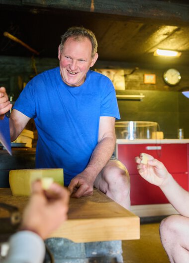 Man in a blue T-shirt holds a note and talks with two other people in a cozy room. | © Kleinwalsertal Tourismus eGen • Photographer: Oliver Farys