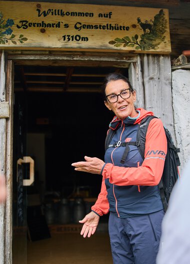 Woman greeting a group of hikers outside the cheese and gemstone hut in Kleinwalsertal, surrounded by mountain scenery. | © Kleinwalsertal Tourismus eGen • Photographer: Oliver Farys