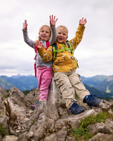 Zwei lachende Kinder auf einer Wanderung in den Bergen, sitzen auf einen Felsen, mit Rucksack und Hands in die Luft gestreckt, fröhlich im Sommer. | © Kleinwalsertal Tourismus eGen • Fotograf: Oliver Farys