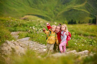 Family with two children hiking on a wooden path in a lush green mountain landscape in Kleinwalsertal. | © Kleinwalsertal Tourismus eGen • Photographer: Oliver Farys