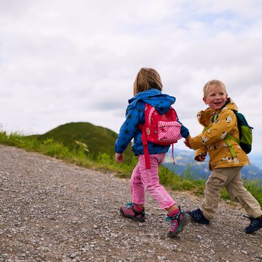 Zwei Kinder wandern auf einem Bergpfad, eines schaut nach hinten und lächelt, getragen von bunten Rucksäcken, im Sommer in den Bergen. | © Kleinwalsertal Tourismus eGen • Fotograf: Oliver Farys
