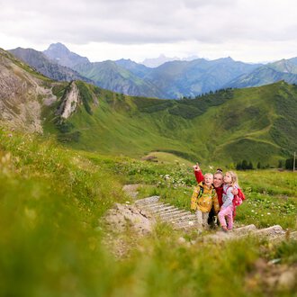 Kinder beim Wandern in den Bergen des Kleinwalsertal, umgeben von grünen Hügeln und hohen Gipfeln. | © Kleinwalsertal Tourismus eGen • Fotograf: Oliver Farys