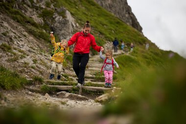 Mutter und zwei Kinder wandern auf einem Bergpfad im Kleinwalsertal, umgeben von grüner Natur, bei schönem Wetter. | © Kleinwalsertal Tourismus eGen • Fotograf: Oliver Farys