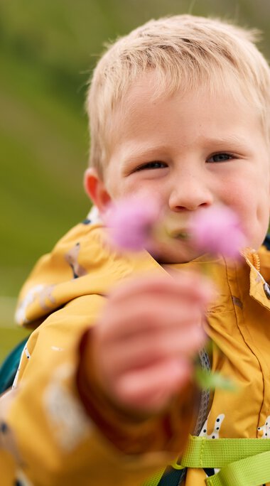 A boy in a yellow rain jacket hiking outdoors, holding a flower towards the camera, with a blurred green landscape in the background. | © Kleinwalsertal Tourismus eGen • Photographer: Oliver Farys