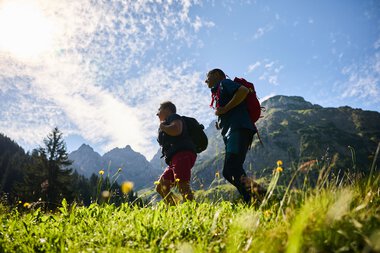 Two hikers with backpacks walking in a mountain landscape during summer, photographed against the sky. | © Kleinwalsertal Tourismus eGen • Photographer: Oliver Farys
