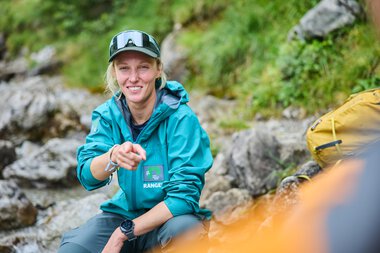 Friendly woman in outdoor clothing, sitting by a river with rocks and nature in the background. | © Kleinwalsertal Tourismus eGen • Photographer: Oliver Farys