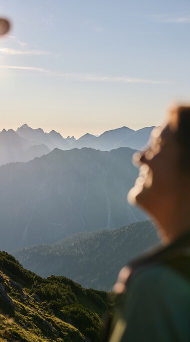 Zwei Wanderer in den Bergen bei Sonnenaufgang im Kleinwalsertal, im Hintergrund die Bergkette, Sonnenlicht hinter einer Person. | © Kleinwalsertal Tourismus eGen • Fotograf: Oliver Farys