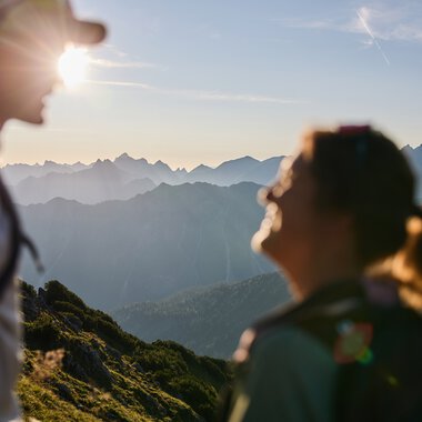 Zwei Wanderer in den Bergen bei Sonnenaufgang im Kleinwalsertal, im Hintergrund die Bergkette, Sonnenlicht hinter einer Person. | © Kleinwalsertal Tourismus eGen • Fotograf: Oliver Farys