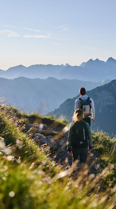 Zwei Wanderer auf einem Bergpfad im Kleinwalsertal mit Blick auf die umliegenden Berge bei Sonnenaufgang. | © Kleinwalsertal Tourismus eGen • Fotograf: Oliver Farys