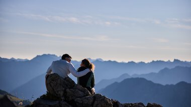 Zwei Menschen sitzen auf einem Berggipfel bei Sonnenaufgang, vor einem Panorama von Bergketten, im Hintergrund der Himmel mit wenigen Wolken. | © Kleinwalsertal Tourismus eGen • Fotograf: Oliver Farys