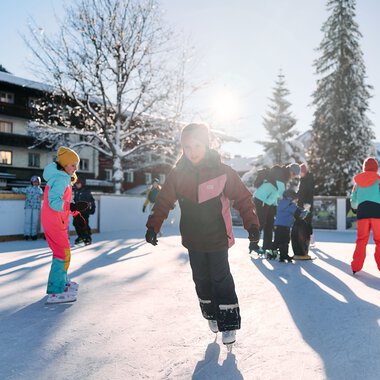 Children ice skating on an outdoor ice rink in winter, surrounded by trees and buildings, sunny day. | © Kleinwalsertal Tourismus eGen • Photographer: Oliver Farys