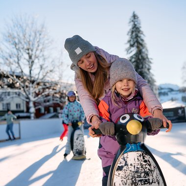 Children skating on an icy rink in Riezlern, Kleinwalsertal, during winter with snow-covered trees and sunlight. | © Kleinwalsertal Tourismus eGen • Photographer: Oliver Farys