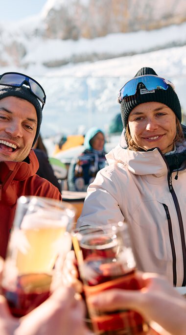 Two smiling people enjoying lunch in the snow, clinking glasses, with snowy landscape and other guests in the background. | © Kleinwalsertal Tourismus eGen • Photographer: Oliver Farys